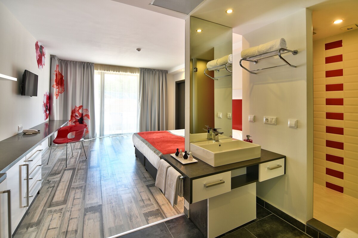 A modern hotel room with a large bed, a bathroom with a sink and mirror, a red chair, and a television on the wall.