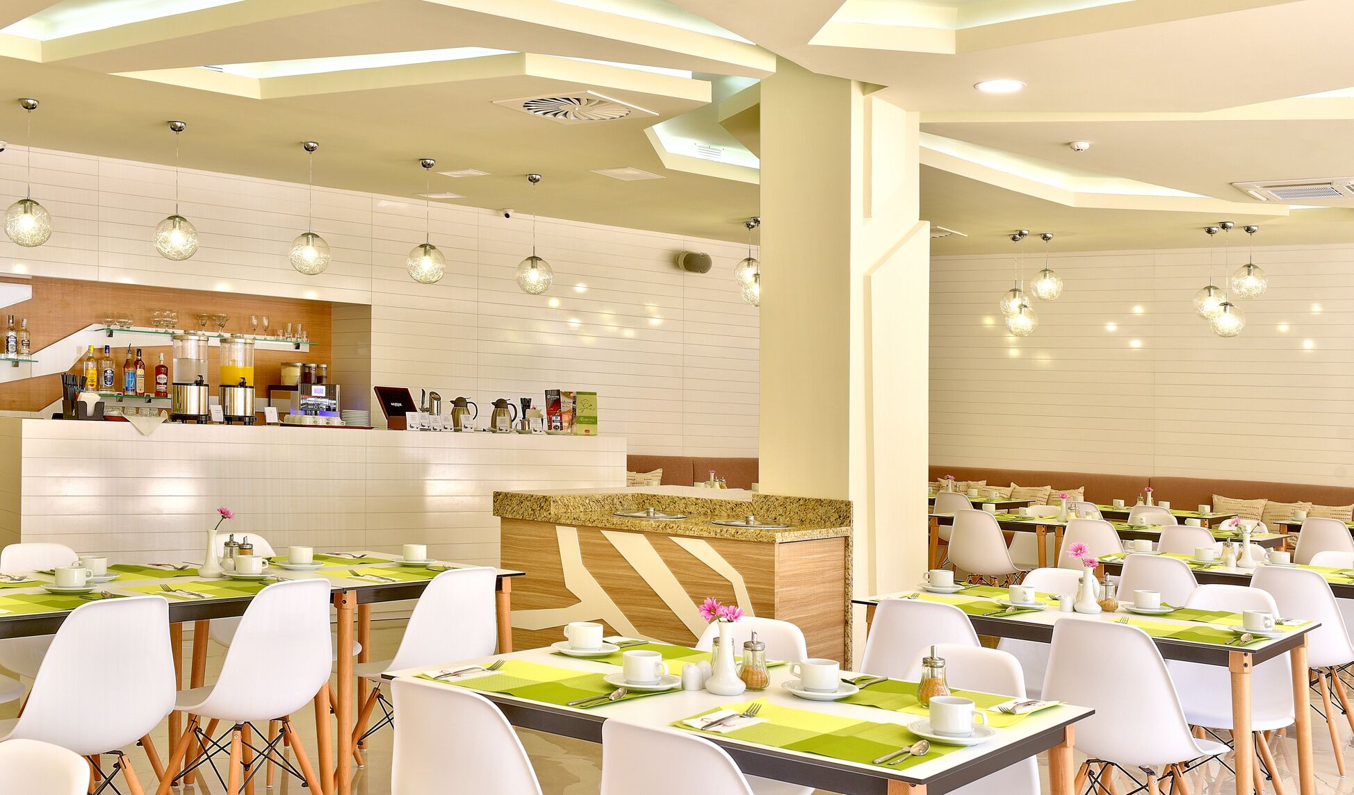 A modern restaurant with a clean, minimalist design, featuring a central counter with a coffee station, multiple dining tables with white chairs, and pendant lighting.