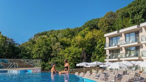 A pool area with lounge chairs and umbrellas, surrounded by lush greenery and a modern building with balconies. - Hotel Grifid Foresta  Adults Only (Golden Sands)