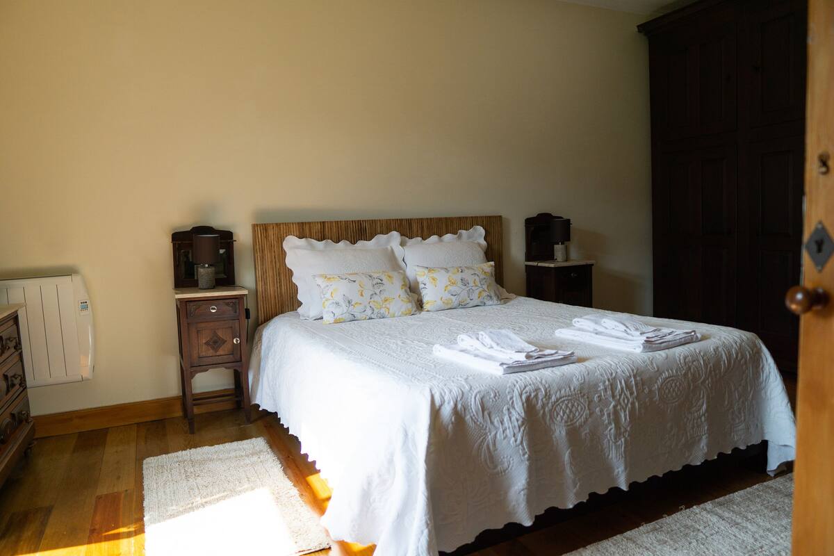 A neatly made bed with white linens and floral pillows, a wooden nightstand with a lamp, and a dark wardrobe.