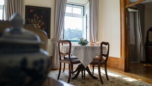 A dining area with a table and chairs, a window with curtains, and a floral painting on the wall. - Casa Mindela Farmhouse (Vila do Conde)