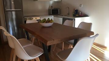 Une cuisine moderne avec une table en bois, des chaises blanches, un saladier de fruits et des appareils électroménagers en acier inoxydable.