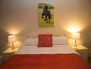 A bedroom with a bed, two bedside lamps, a red pillow, and a painting of a horse on the wall. - Greenbank Farmhouse (Grange-over-Sands)