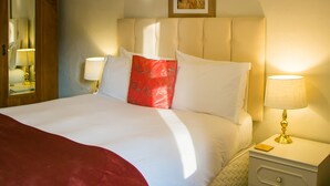A neatly made bed with white linens and a red accent pillow, a bedside lamp, a framed picture on the wall, and a wooden wardrobe.