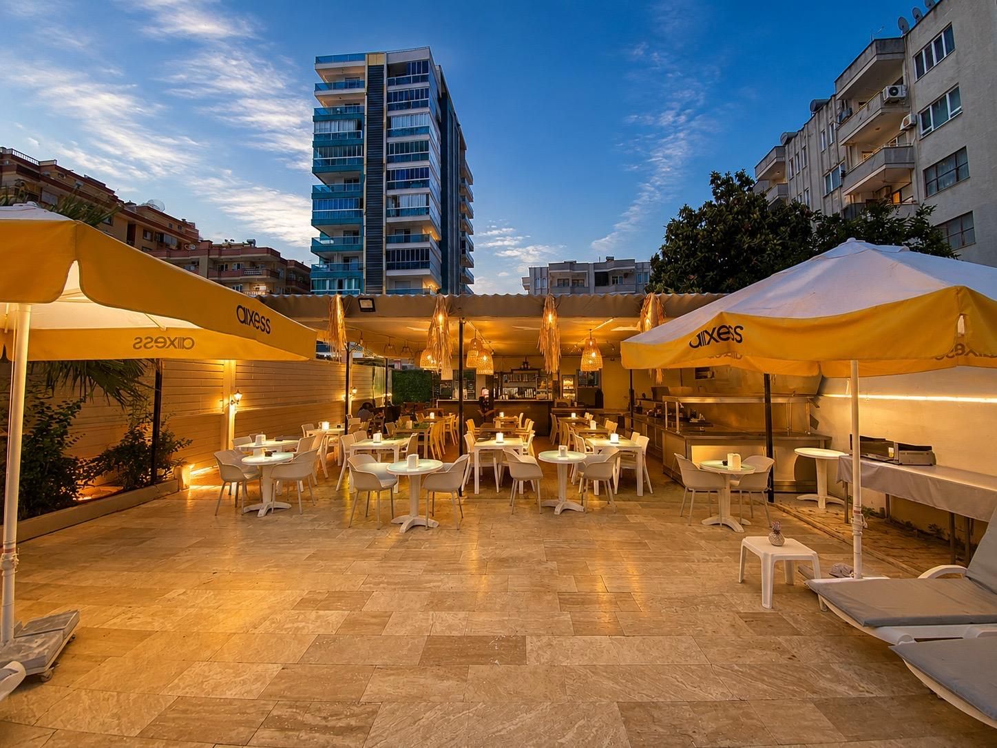 Un espacio de comedor al aire libre con mesas, sillas, sombrillas y vistas a un edificio alto al fondo.