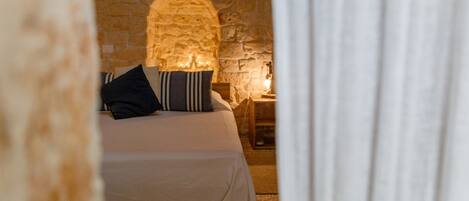 A cozy room with a stone wall, a bed with a white bedspread and striped pillows, and a nightstand with a lamp.