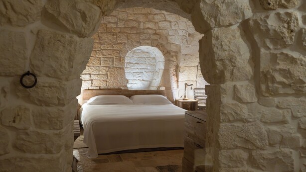 Une chambre aux murs de pierre, avec un lit, une table de chevet en bois et une fenêtre ronde.