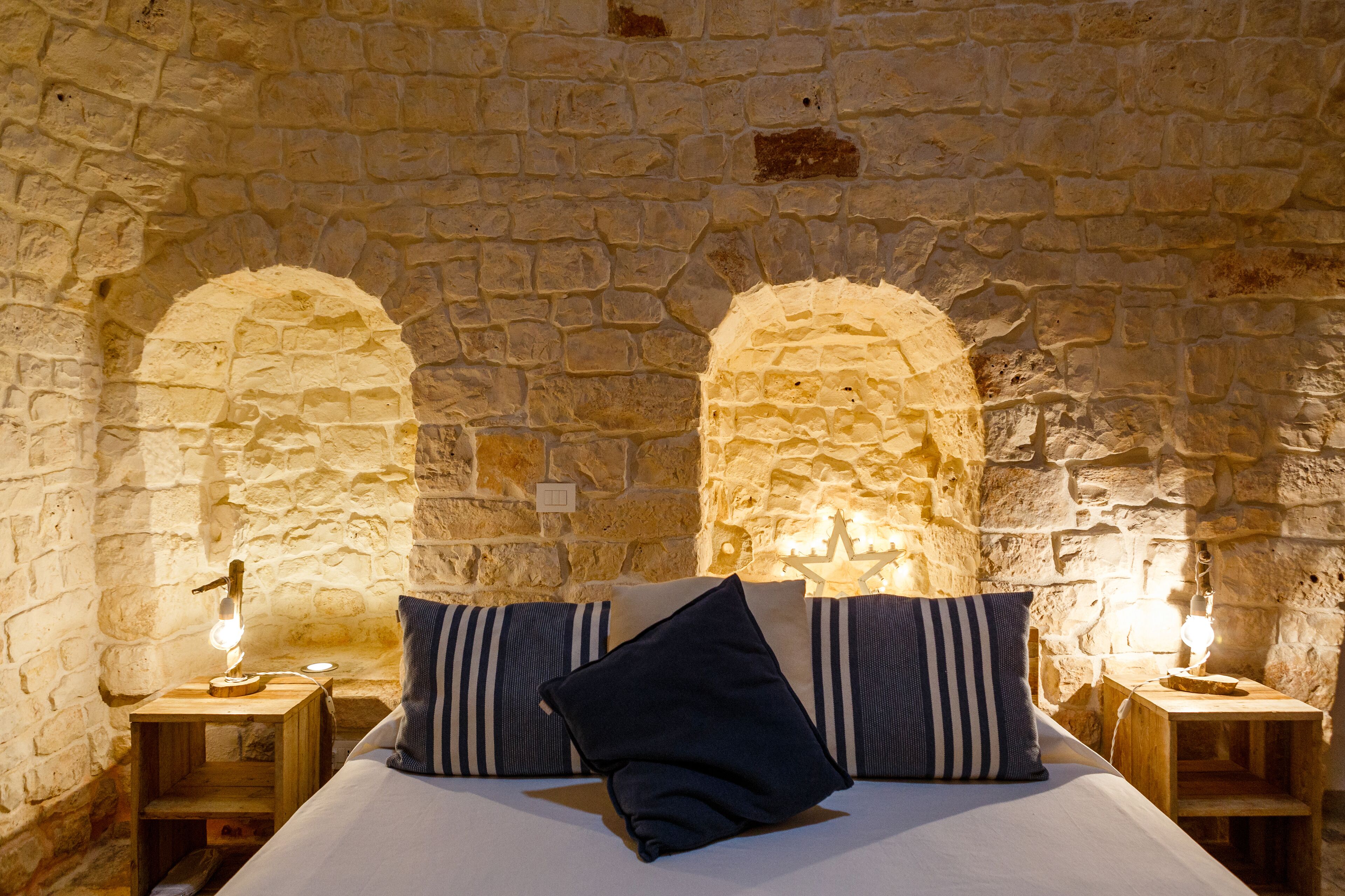 A bedroom with a stone wall, two nightstands, a bed with striped pillows, and a lit wall lamp.