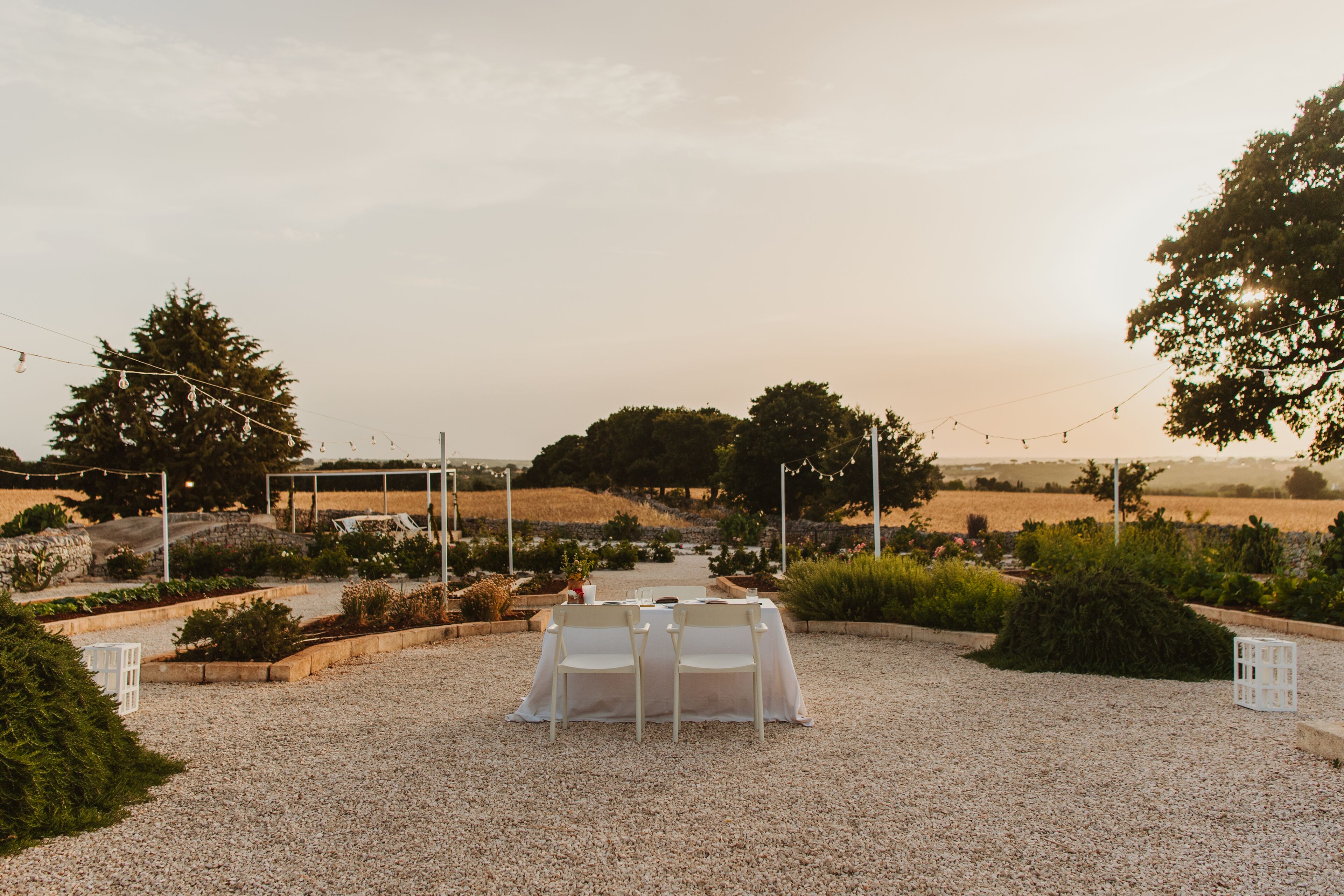 Montagem de casamento com mesa e cadeiras em área de cascalho, cercada por vegetação e árvores.