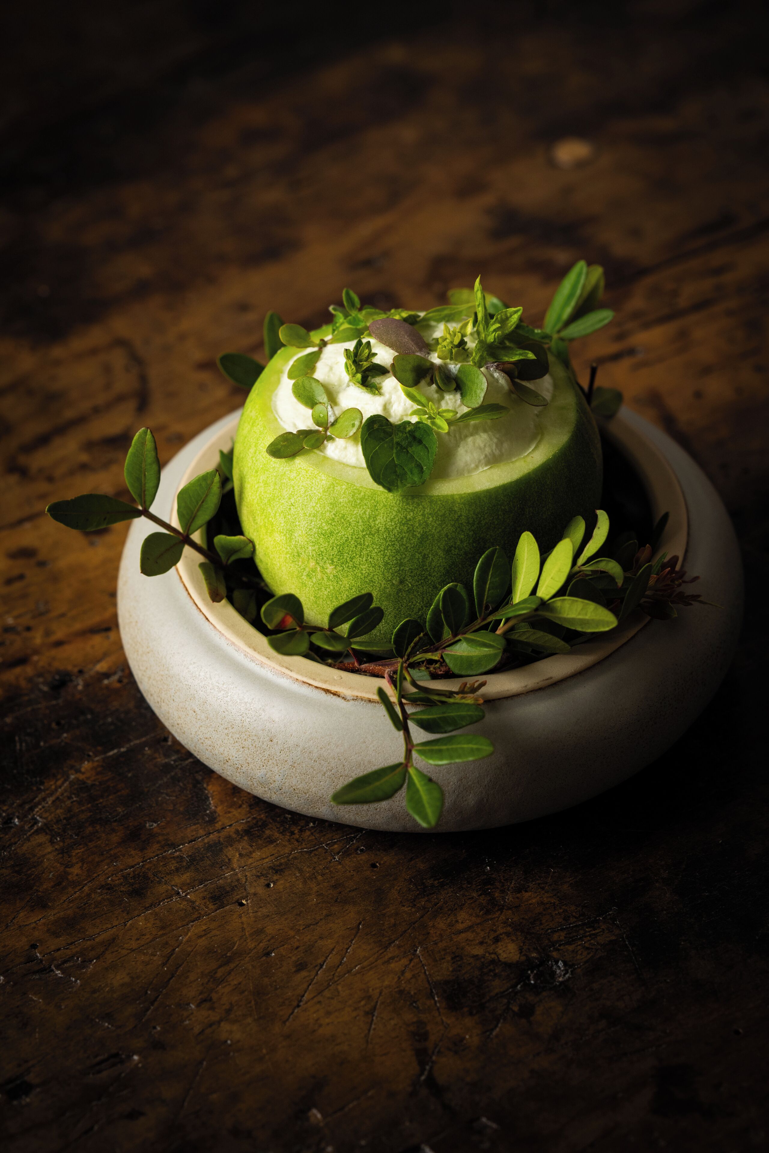 A green fruit sculpture with leaves and a white topping, placed on a round ceramic plate on a wooden surface.