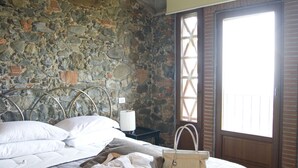 Slaapkamer met een stenen muur, een bed met een grijze dekbedovertrek, een hangende lamp en een raam met houten luiken.