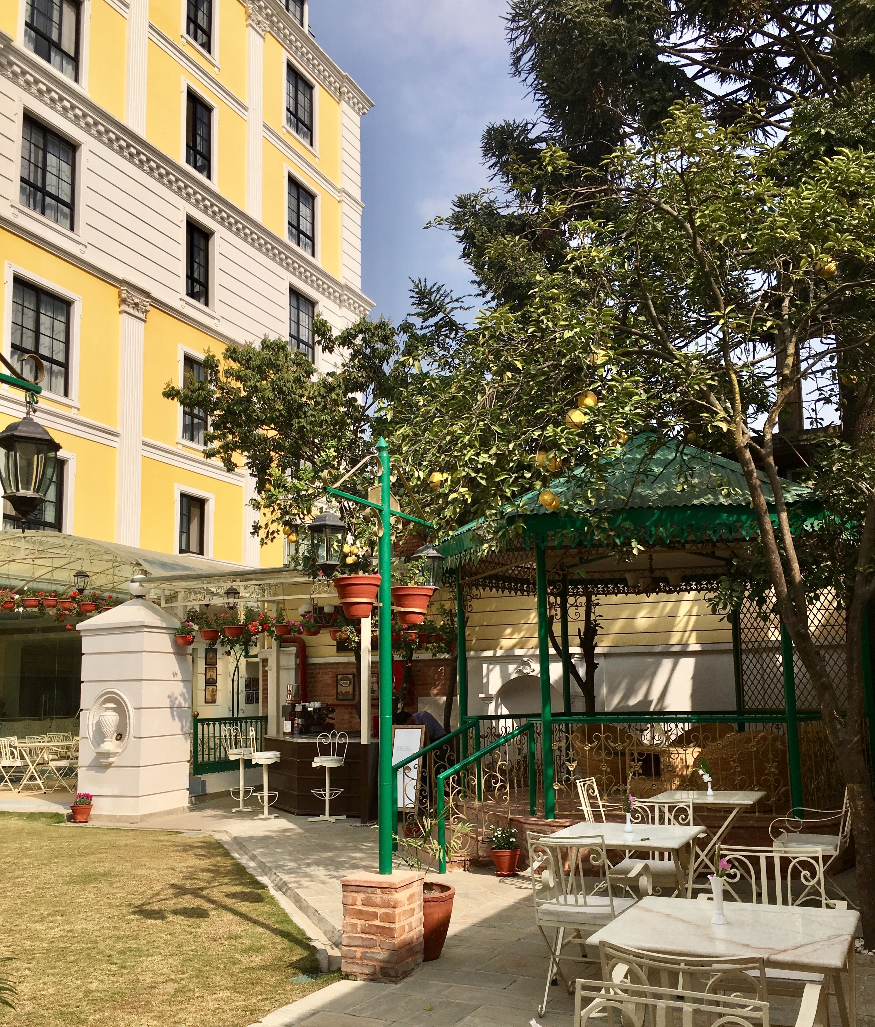 An outdoor seating area with tables and chairs, a green pergola, and a building with yellow and white exterior walls in the background.