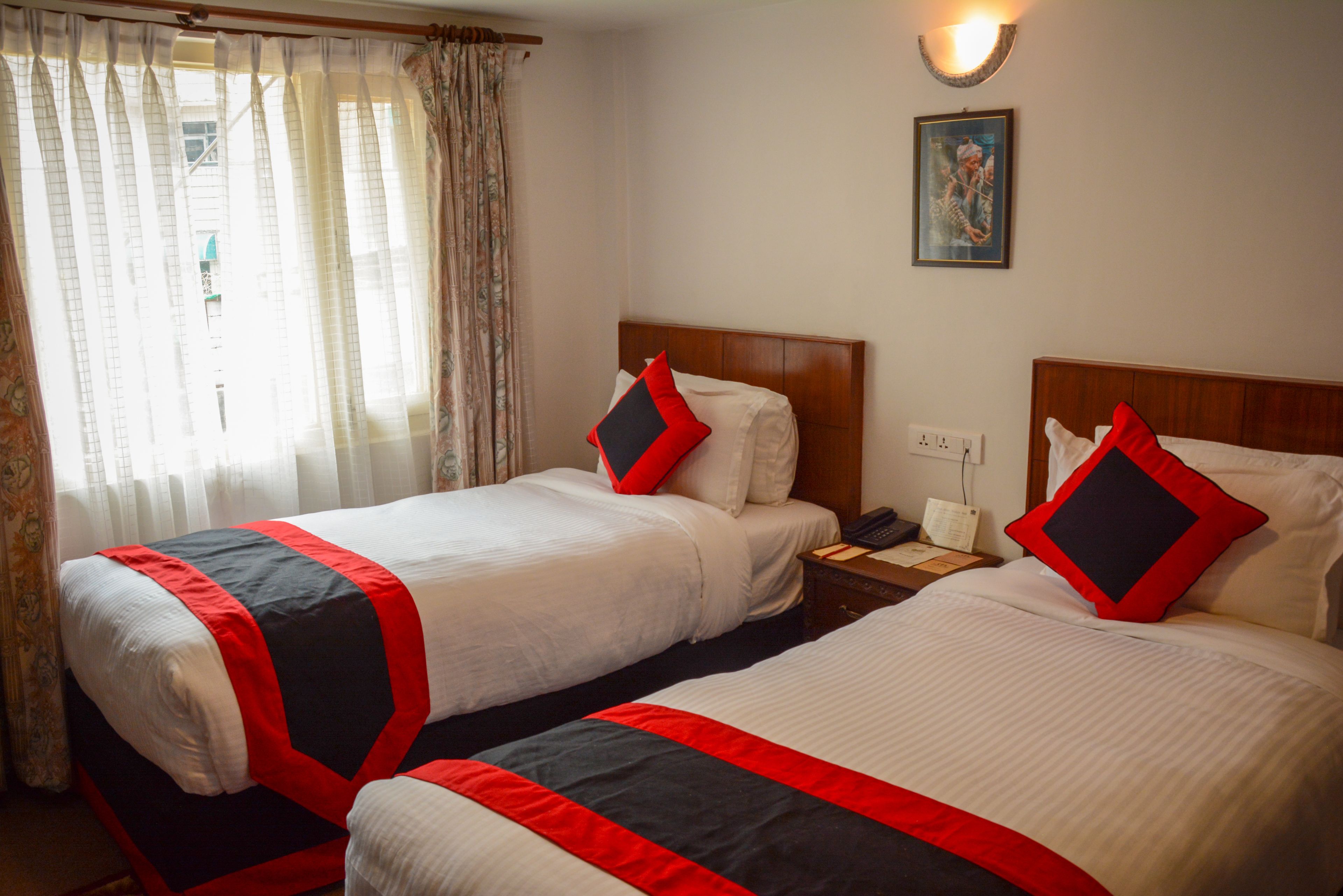 A hotel room with two beds, each with a red and black bedspread, white pillows, and a wooden headboard. There is a window with sheer curtains, a wall-mounted light fixture, and a painting above the beds.