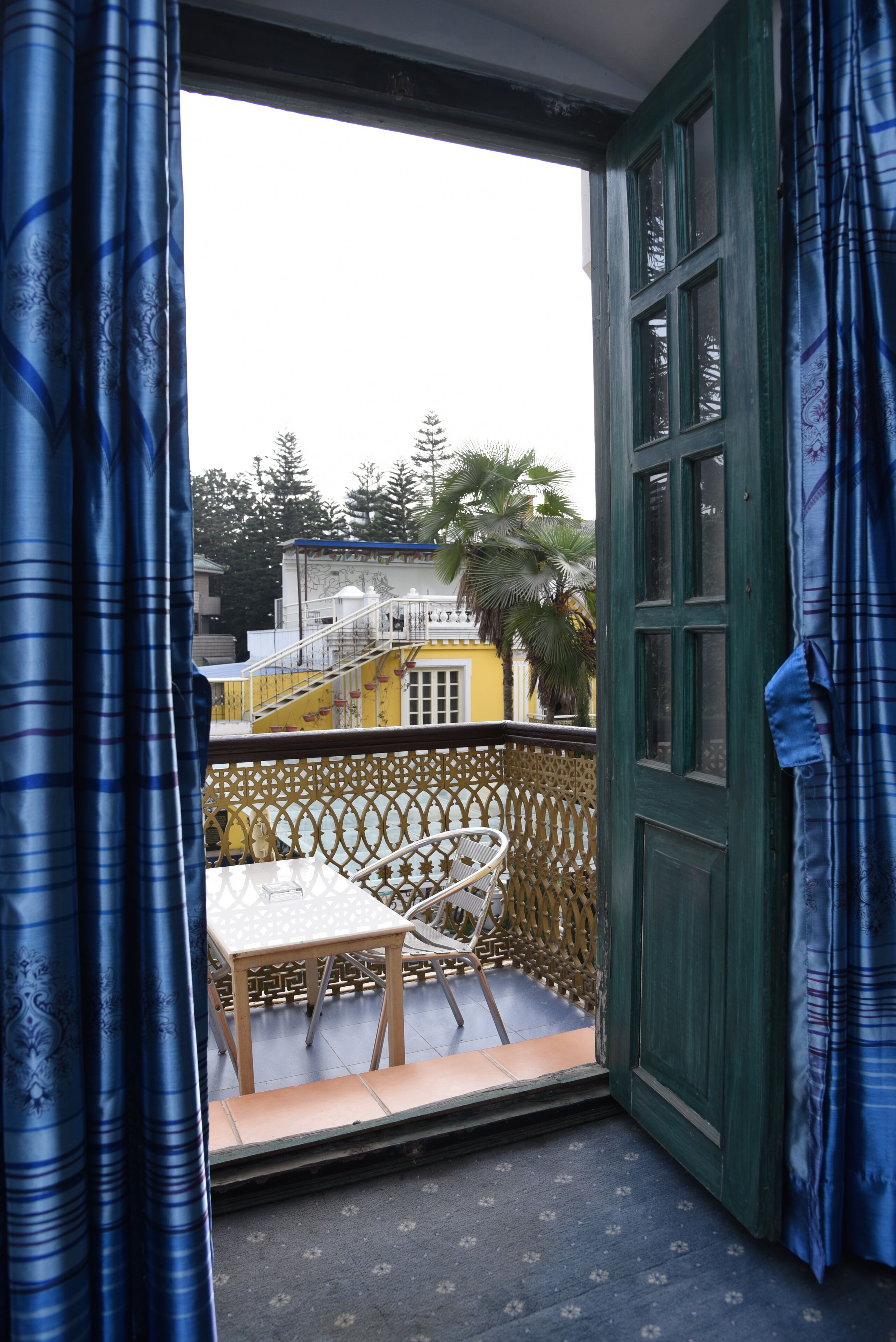 Un balcón con una mesa y silla de madera, una puerta verde y vista a un edificio amarillo con una escalera.