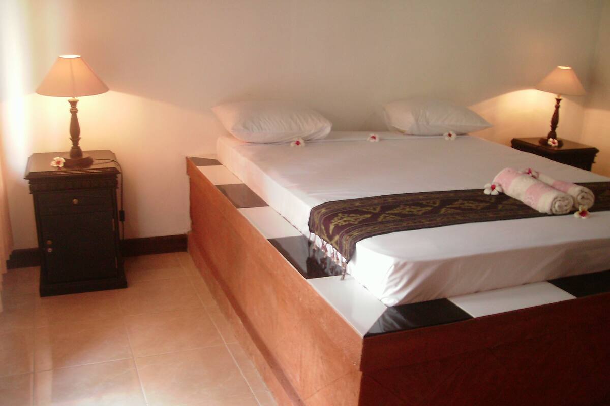 A neatly made bed with a white comforter and a patterned blanket, flanked by two bedside tables with lamps, in a room with tiled flooring.