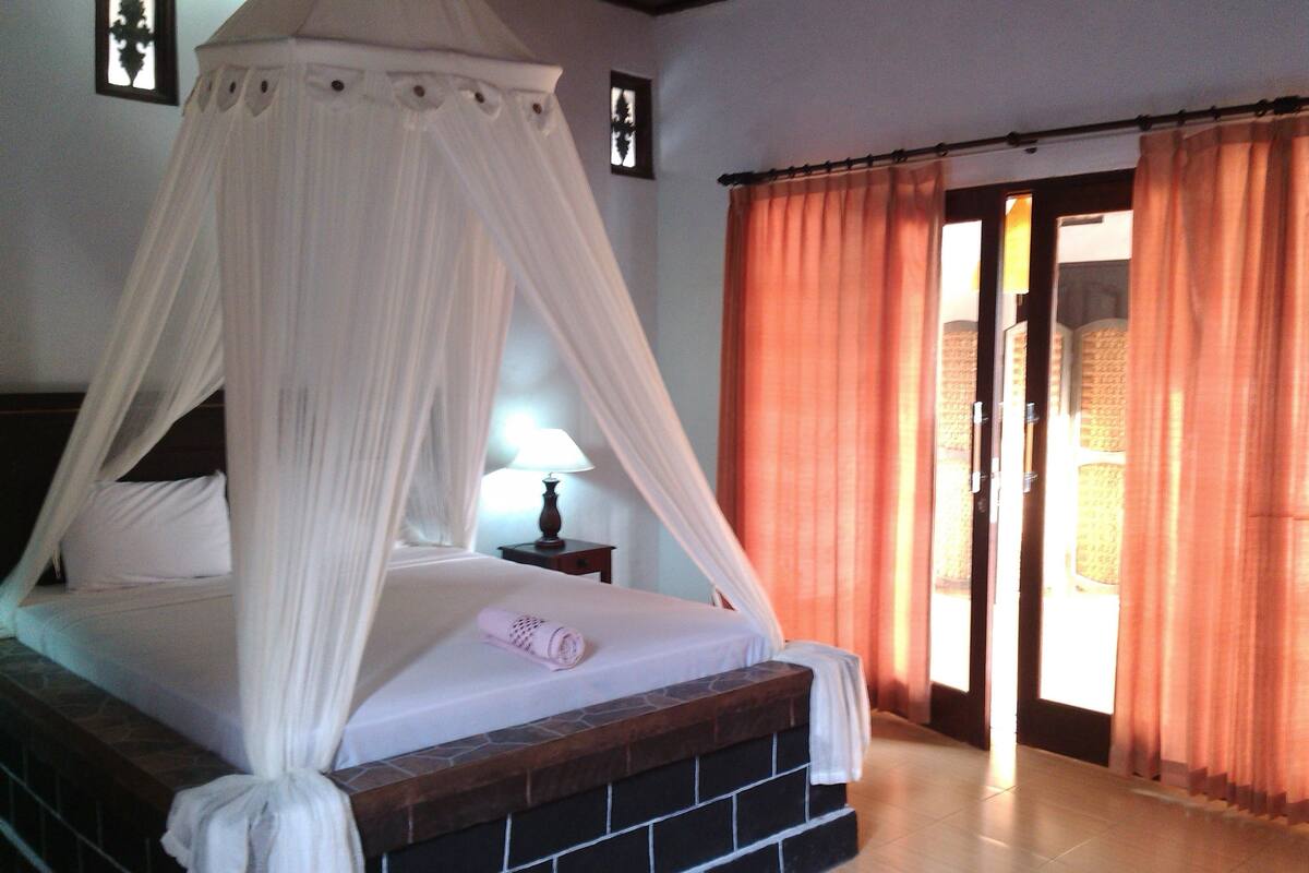 A bedroom with a four-poster bed, a nightstand with a lamp, and a view of the outside through an open door.