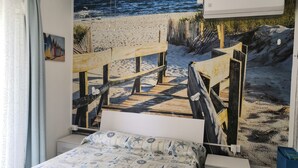 Un dormitorio con un amplio mural de una playa, una cama con colcha estampada, un aparador blanco y un baúl azul.