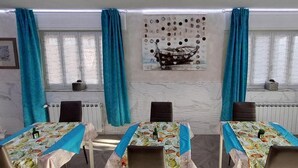 Un comedor con mesas dispuestas para comidas, cortinas azules y un elemento decorativo sobre las ventanas.