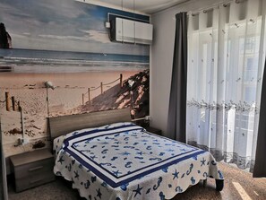 A bedroom with a bed, a beach mural on the wall, a window with curtains, and a small bedside table. - Villa La Lanterna (Pietra Ligure)