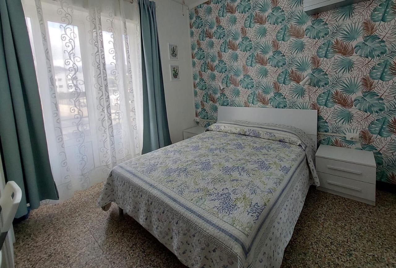 Une chambre à coucher avec un lit, une table de chevet, une fenêtre avec des rideaux et un mur orné d’un motif de feuilles.