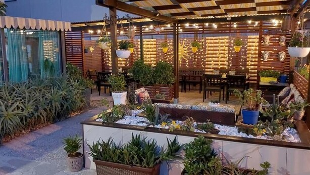 Un patio al aire libre con mesas y sillas de madera, rodeado de plantas en macetas y un muro de ladrillo.