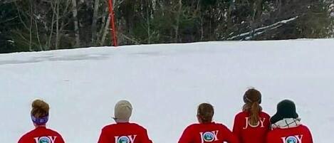 Vier Personen sitzen auf Schneebrütten, auf denen 'JOY' steht, in einer verschneiten Winterlandschaft.