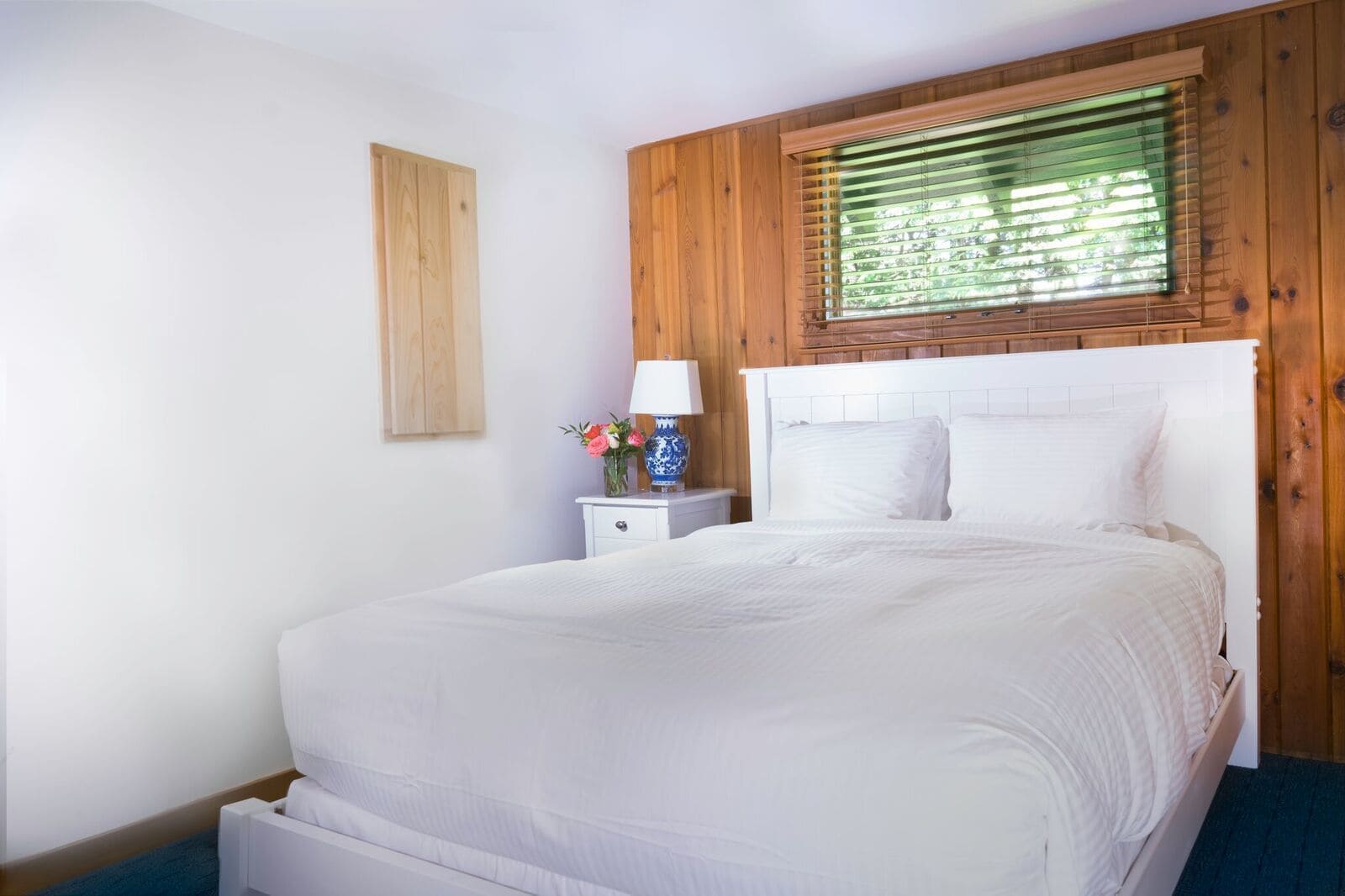 Un dormitorio con cabecera de madera, ropa de cama blanca, una mesita de noche con lámpara y un jarrón con flores.