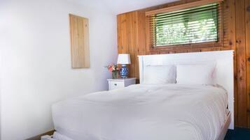 Un dormitorio con cabecera de madera, ropa de cama blanca, una mesita de noche con lámpara y un jarrón con flores.
