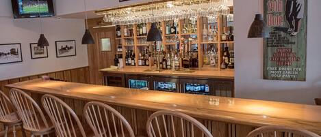 Una zona de bar con mostrador de madera, varios taburetes y una variedad de botellas y vasos en estantes.