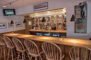 A bar area with wooden bar counter, multiple stools, and a variety of bottles and glasses on shelves. - Leland Lodge (Leland)