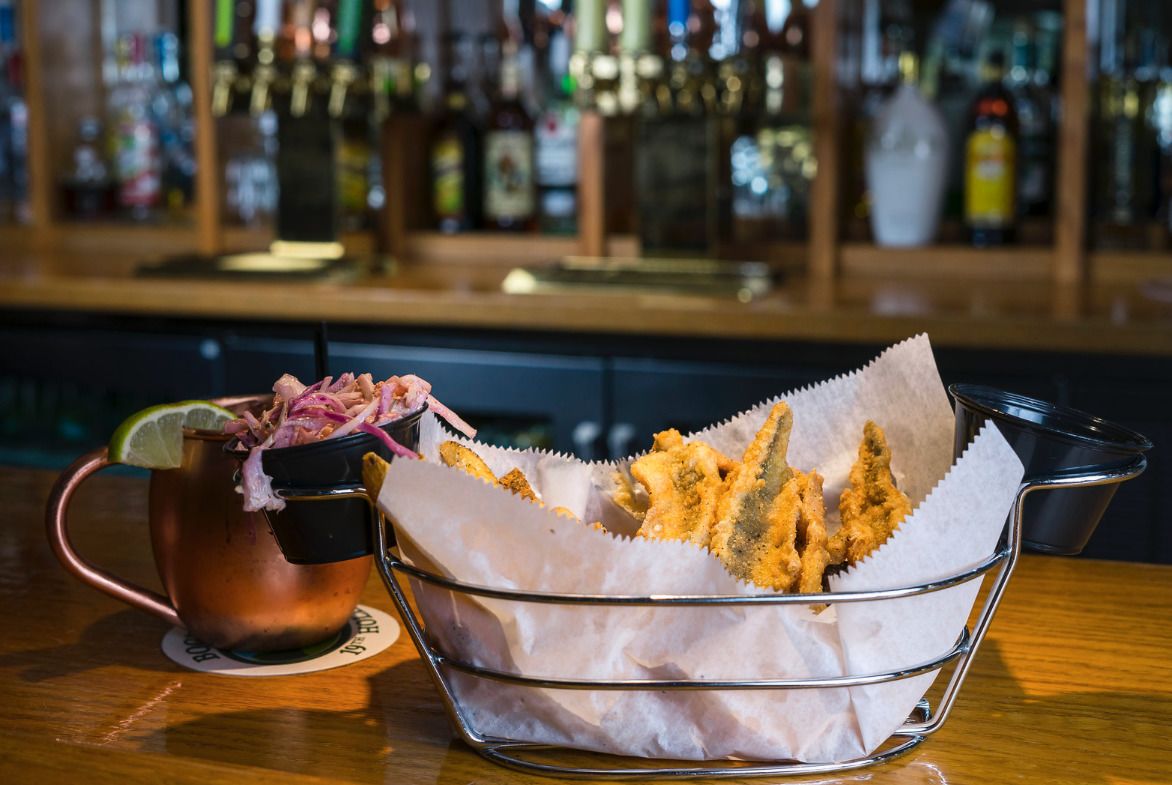 Una taza de cobre con un gajo de lima, una cesta de pescado frito y una barra con varias botellas al fondo.