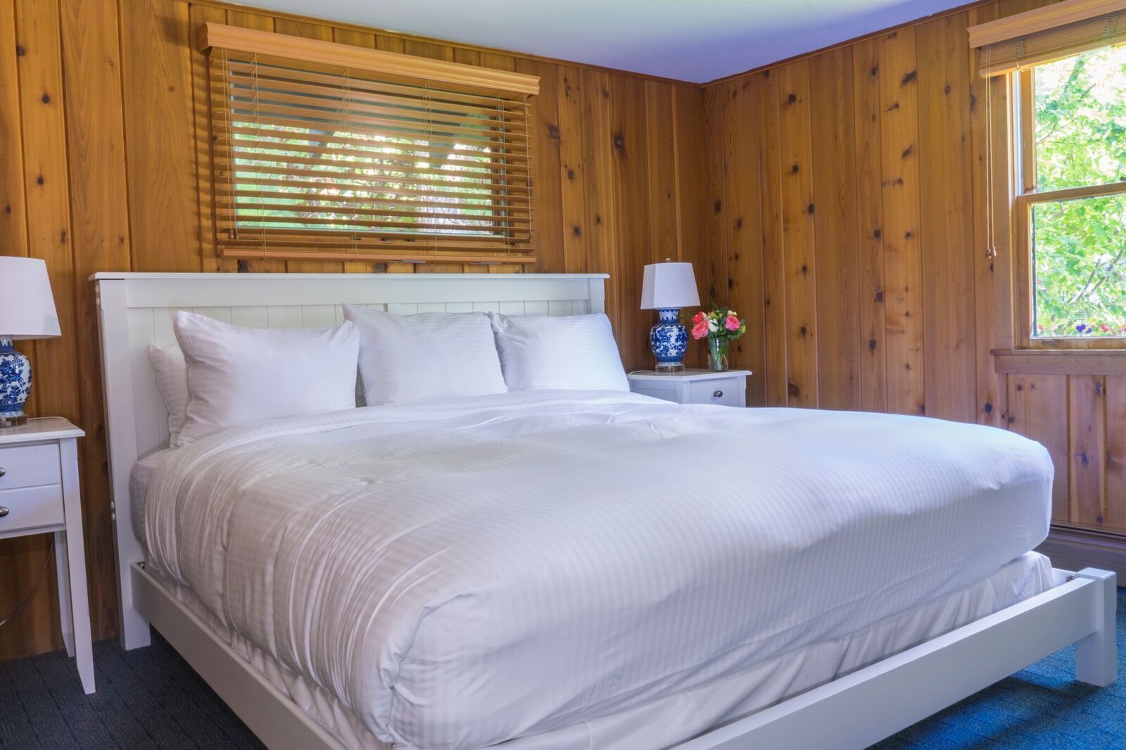A large bed with white linens in a room with wooden walls and a window with blinds.