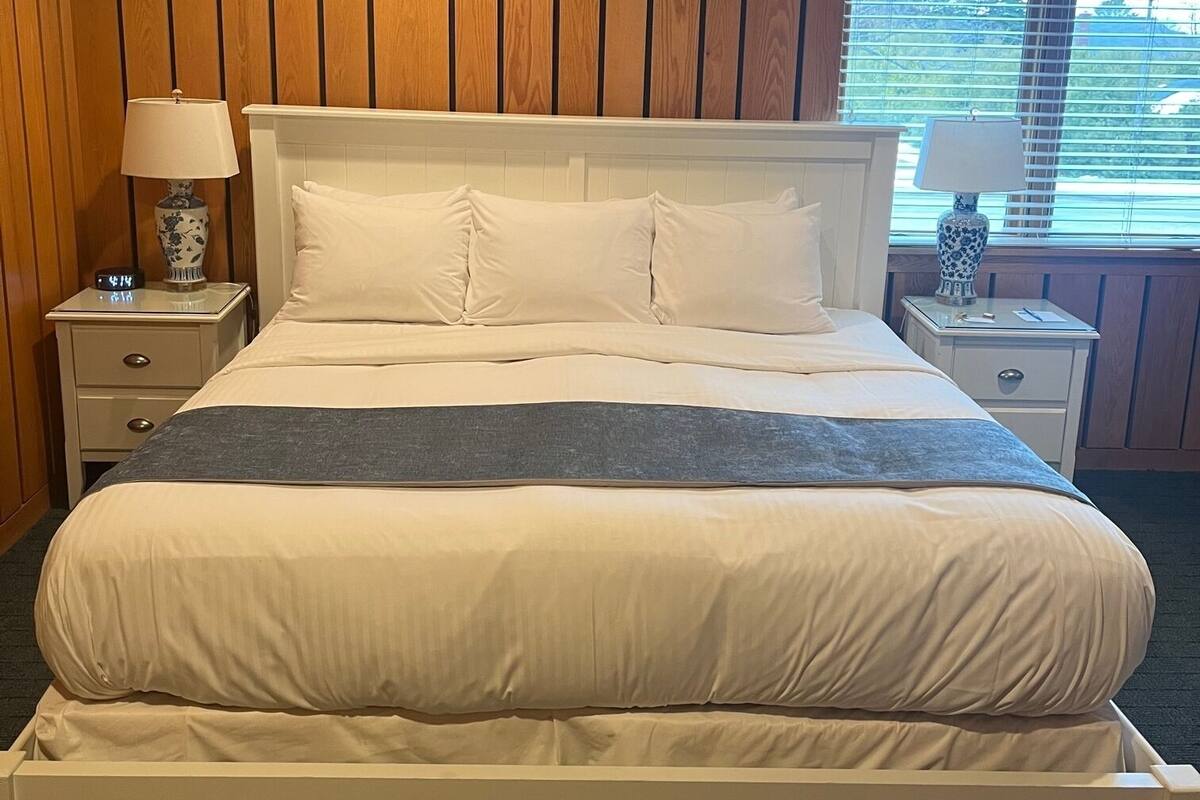 A neatly made bed with white bedding and a gray throw, flanked by two bedside tables with lamps, in a room with wooden paneling and a window with blinds.