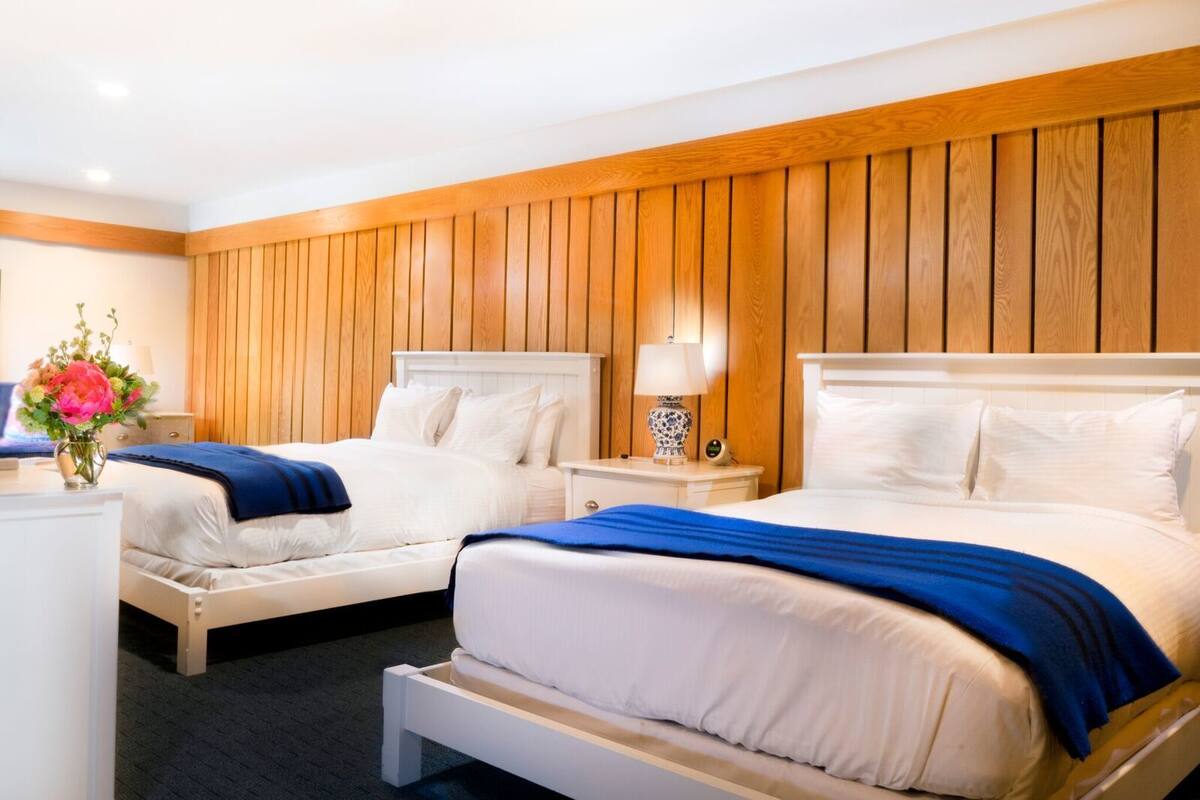 A hotel room with two single beds, wooden paneling on the walls, a nightstand with a lamp, and a vase with flowers.