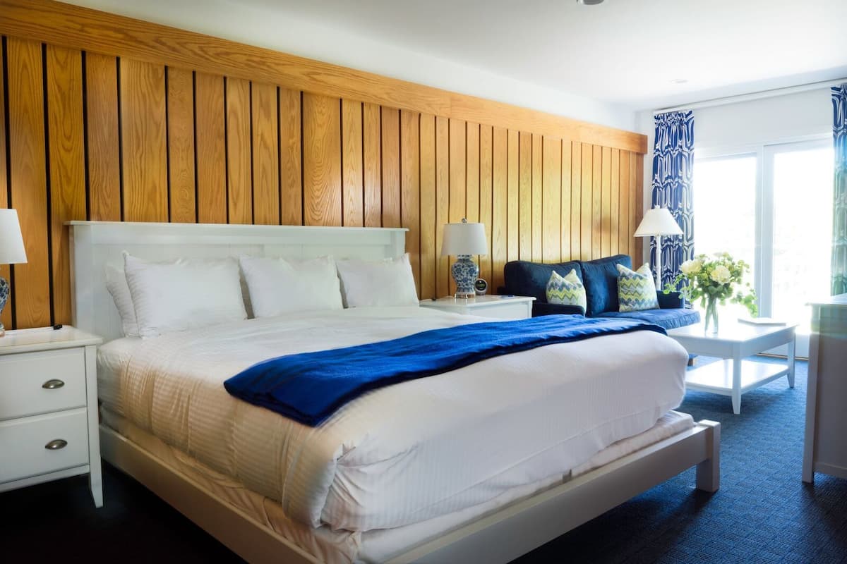 A hotel room with a large bed, wooden panel walls, a bedside table with a lamp, a small sofa with cushions, and a view of the outdoors through a window.