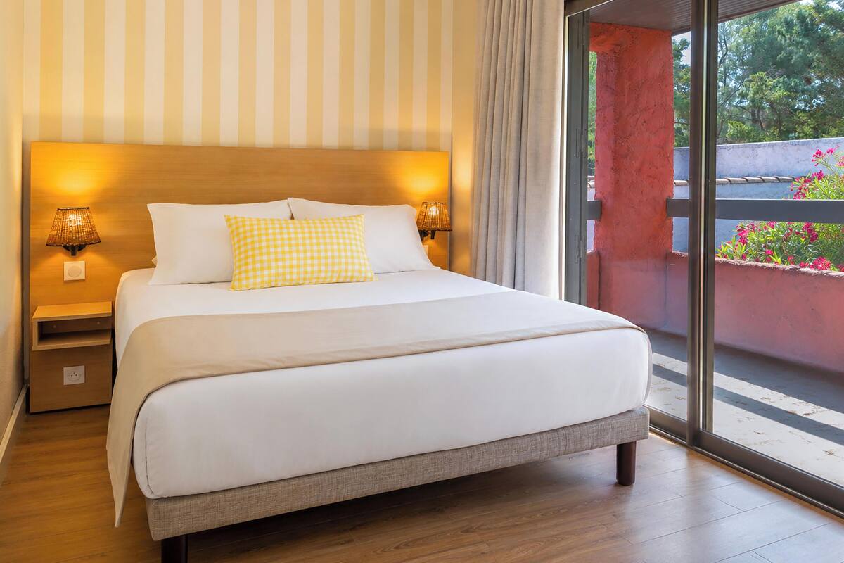 Ein Bett mit weißer Bettwäsche und einem karierten Kopfkissen, zwei Nachttischlampen, ein Nachttisch und der Blick auf einen Balkon mit Pflanzen.