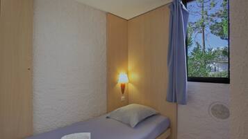 Une petite chambre avec un lit simple, une tête de lit en bois, une lampe de chevet et une fenêtre donnant sur des arbres et une maison.