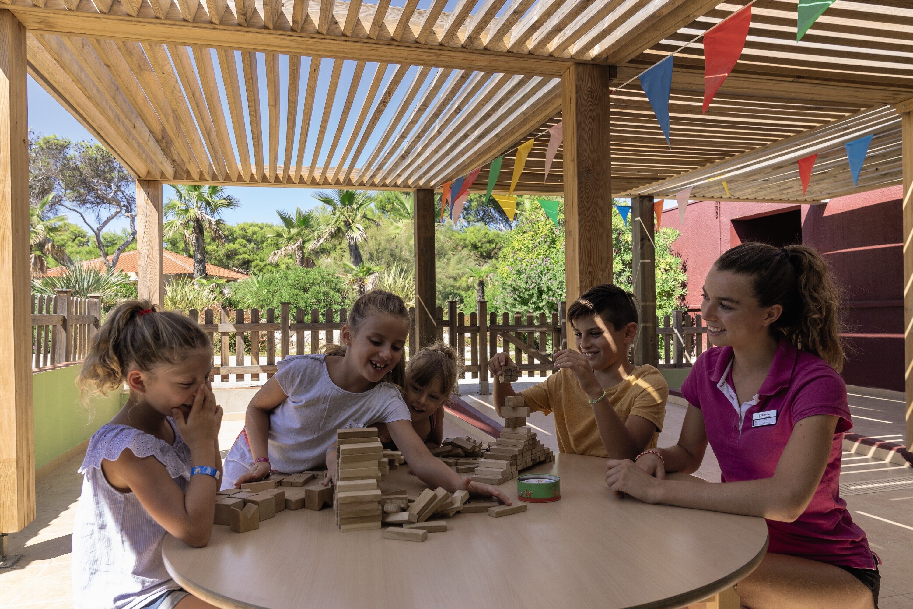 A group of people playing with wooden blocks at a wooden table under a pergola with colorful flags.