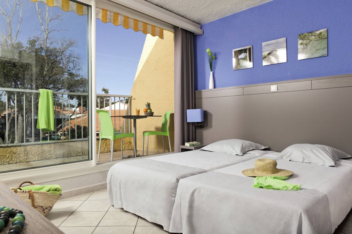 Quarto de hotel com duas camas, varanda com mesa e cadeiras, e televisão na parede.