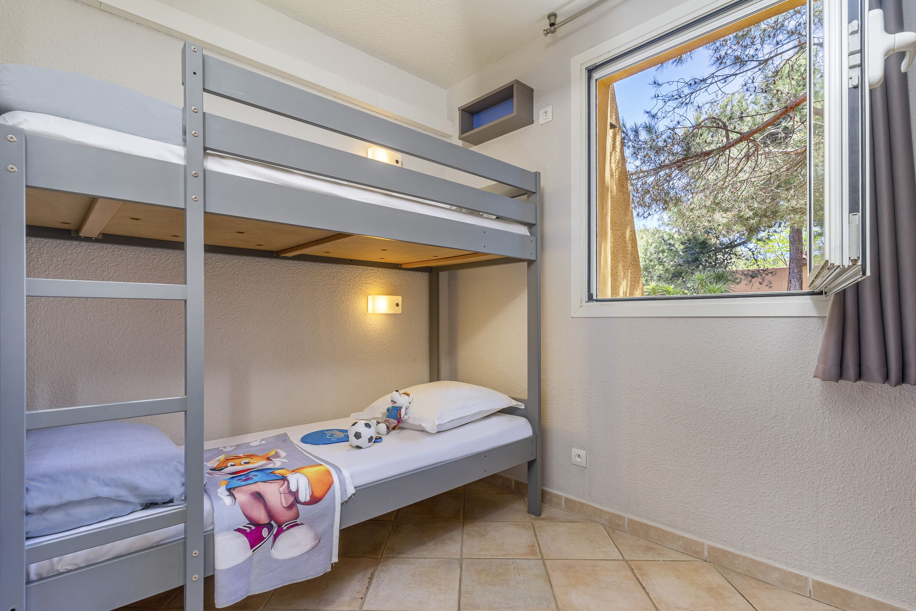 A bunk bed room with a window, a shelf above the upper bunk, and a small window with curtains.