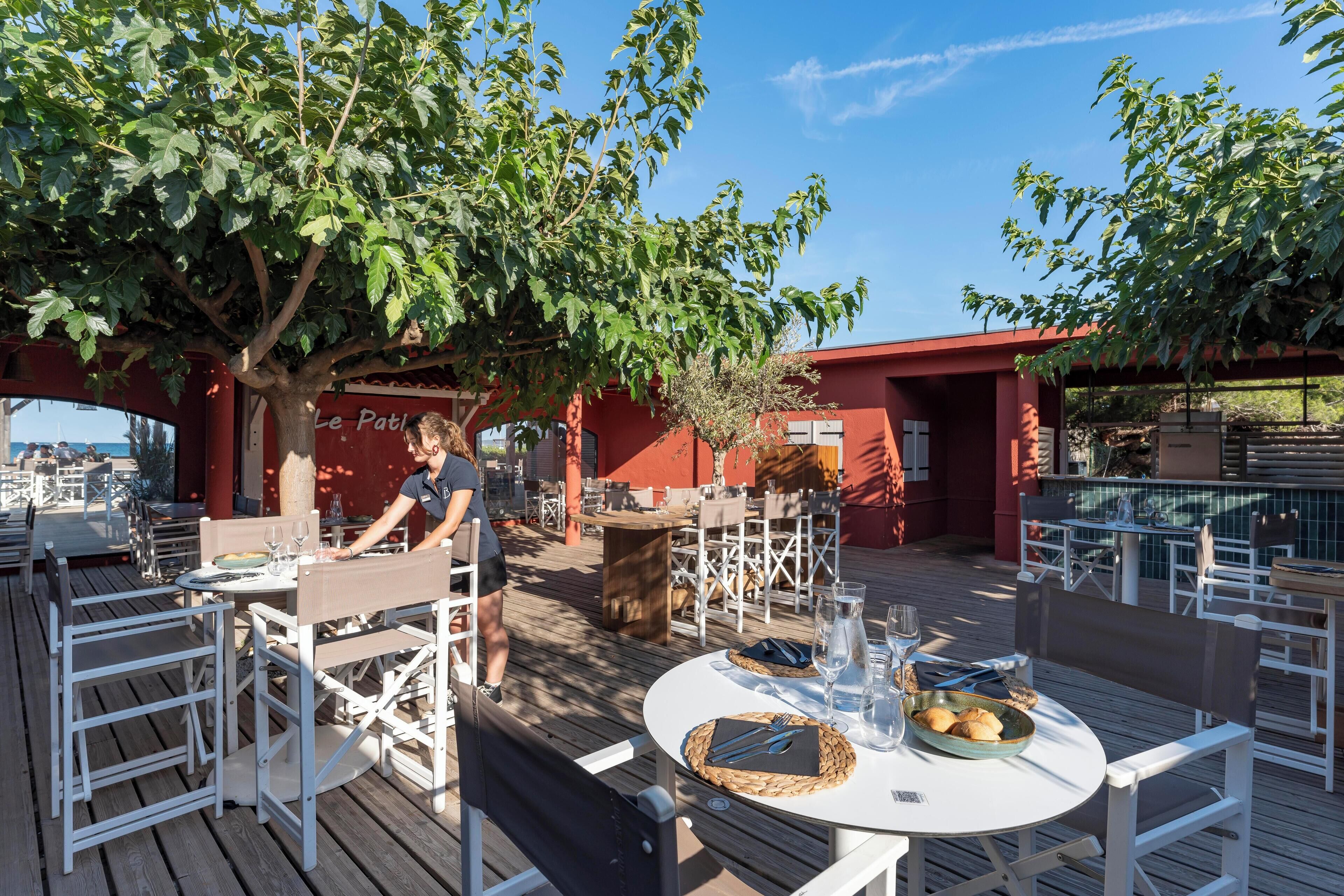 Un espace extérieur aménagé pour les repas, avec des tables et des chaises blanches, un arbre qui offre de l’ombre et un bâtiment rouge en arrière-plan.