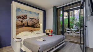 A bedroom with a bed, a nightstand, a reading lamp, a window with a view of an outdoor seating area. - Belambra Clubs Presqu'île de Giens - Riviera Beach Club (Hyères)