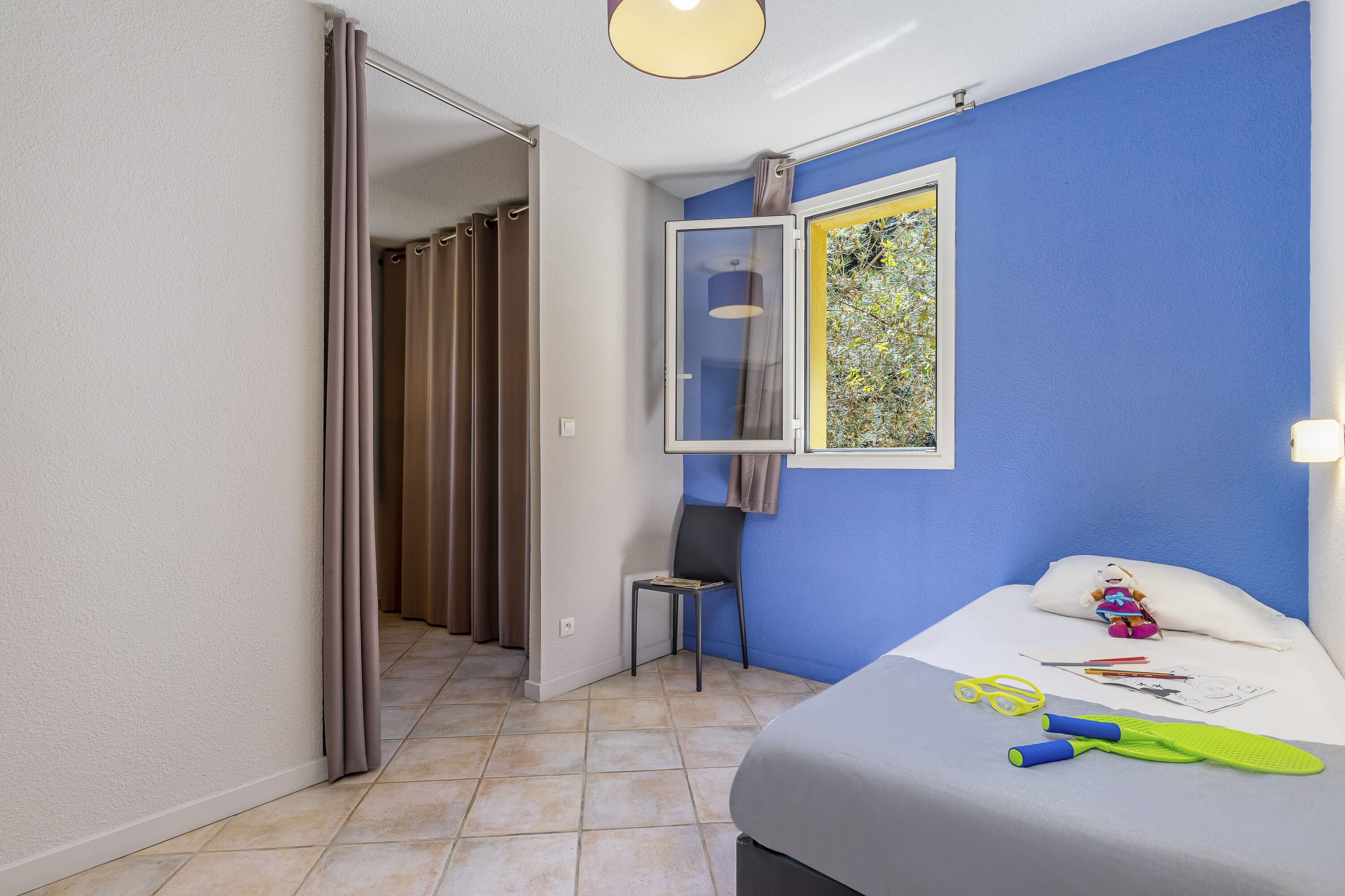 A bedroom with a bed, a chair, a window with curtains, and a wall-mounted lamp.
