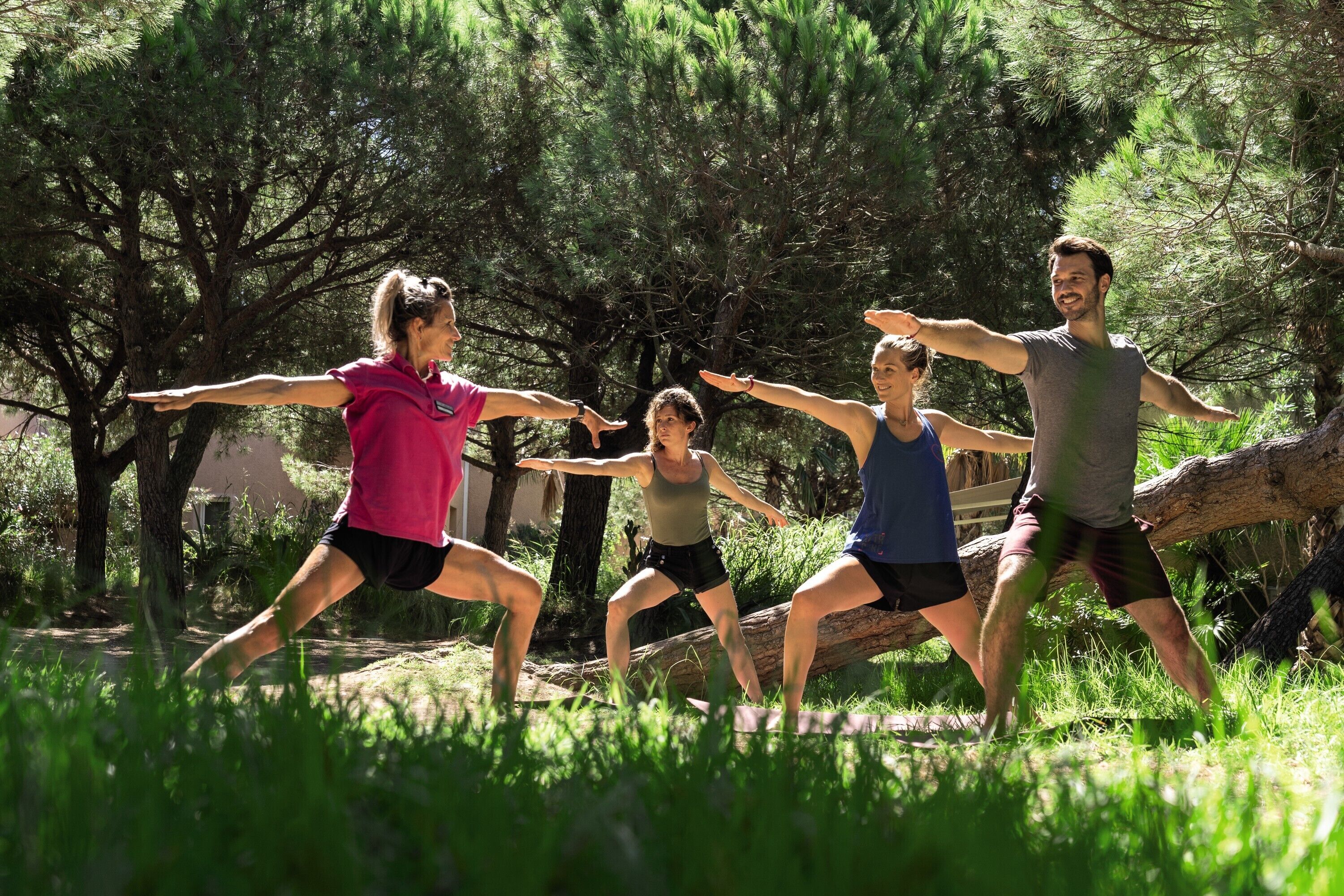 A group of people performing a stretching exercise outdoors in a natural setting with trees and a log in the background.