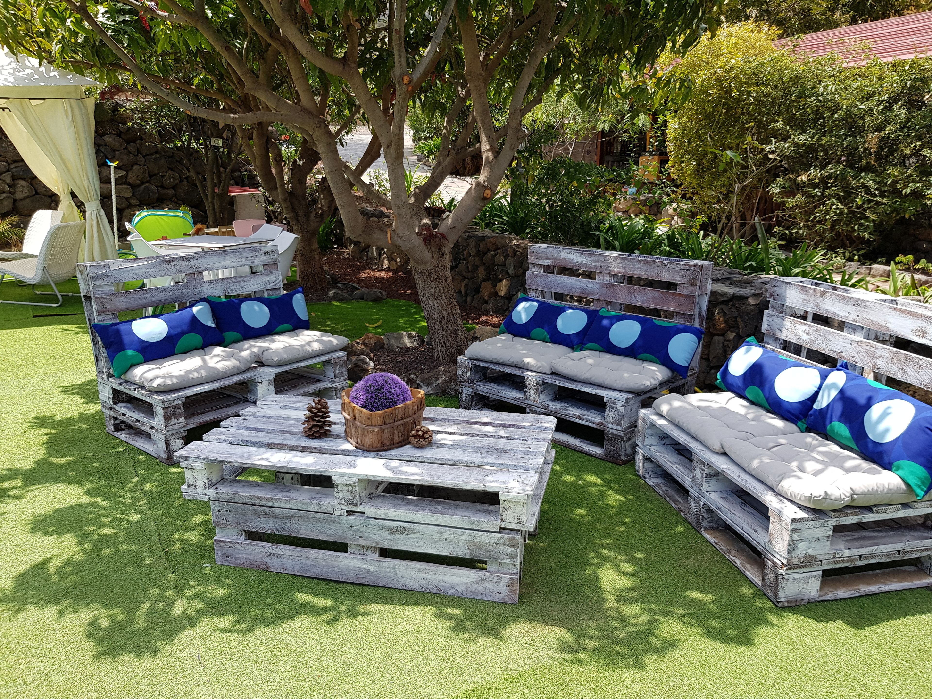 Un espace extérieur aménagé dans un style rustique, avec des bancs en bois et des coussins, entouré de verdure et d’un arbre.