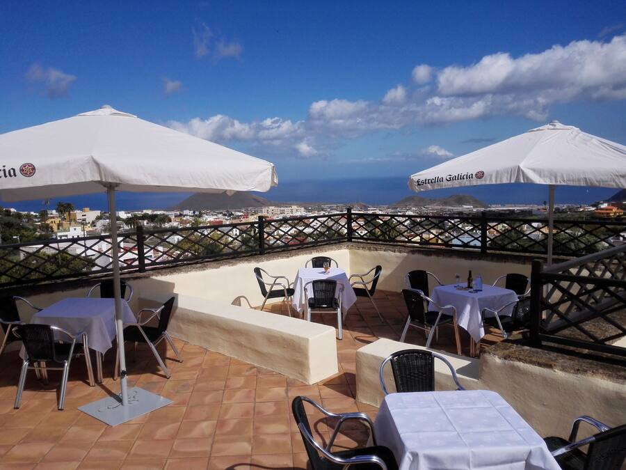 Una terraza al aire libre con mesas y sillas, sombrillas blancas y vistas a la ciudad y al mar.