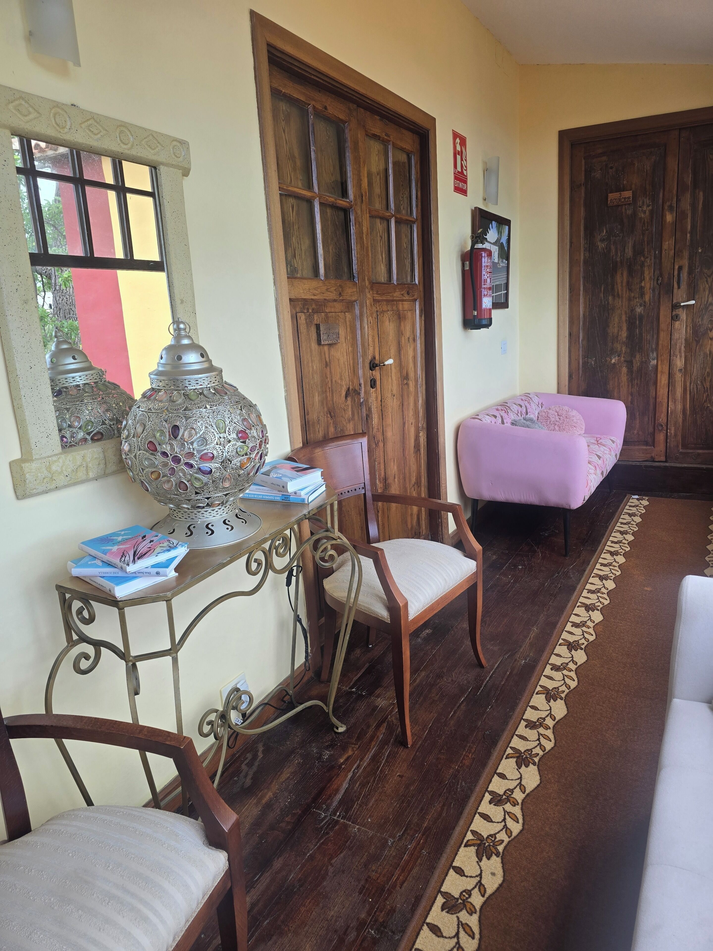 A room with a wooden door, a table with books, a chair, and a pink ottoman.