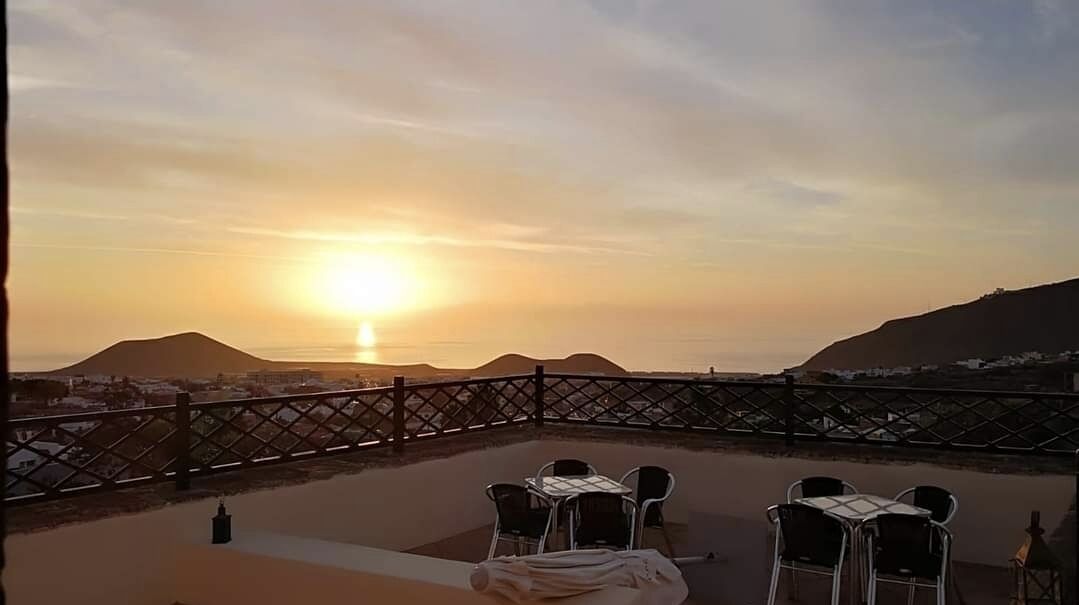Una terraza con vistas a la puesta de sol sobre montañas, con una mesa y sillas.
