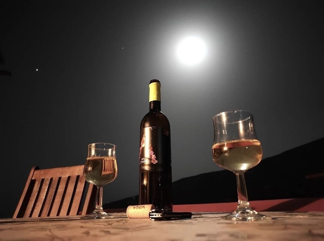 A bottle of wine, two glasses, and a corkscrew on a table with a wooden bench in the background, under a night sky with a bright moon.