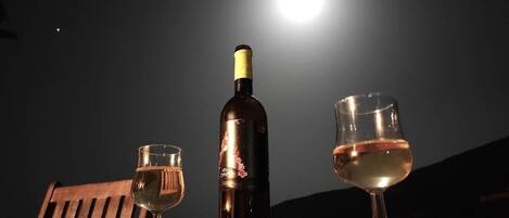 A bottle of wine, two glasses, and a corkscrew on a table with a wooden bench in the background, under a night sky with a bright moon.